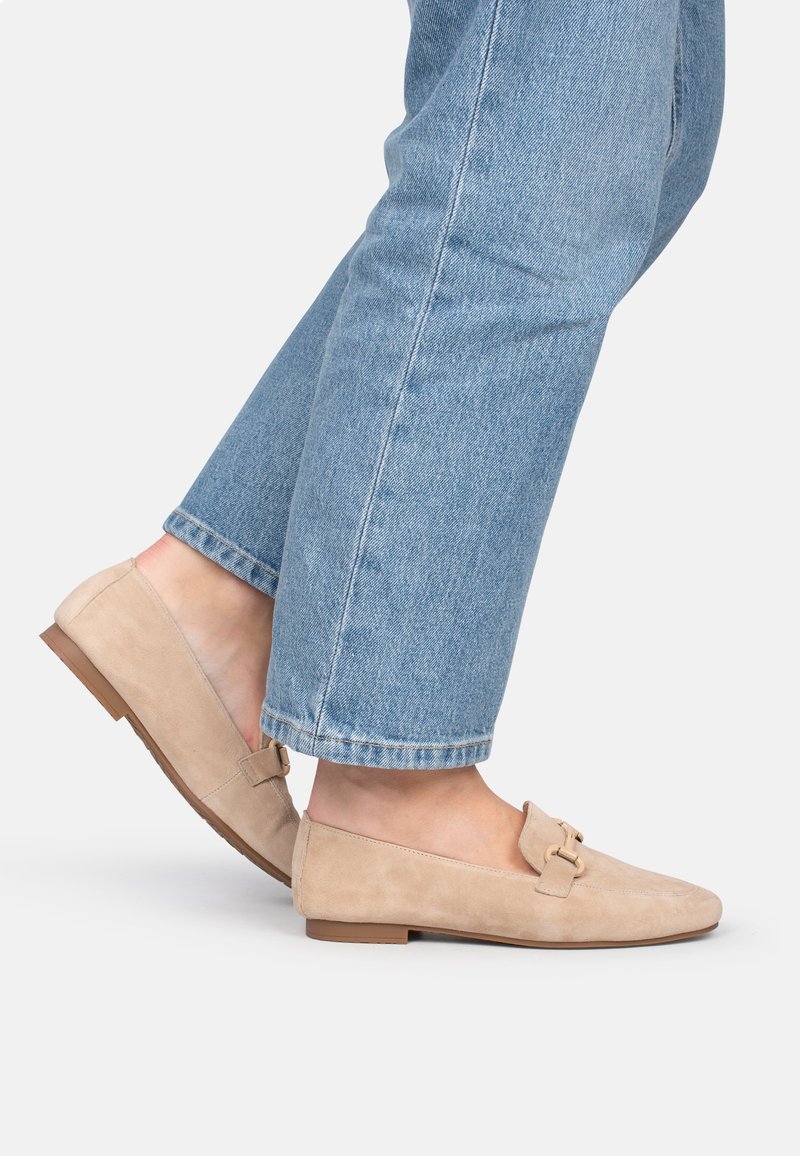 Mocasines de ante beige con tacón plano, que cuentan con un detalle decorativo de hebilla dorada y puntera redonda, combinados con unos jeans de longitud hasta el tobillo de color azul claro.