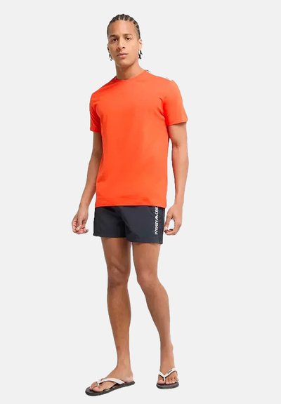 Camiseta de manga corta de color naranja brillante con un diseño simple, combinada con pantalones cortos de baño oscuros y chanclas; tejido suave y ajuste casual.