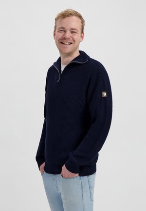 Dunkelblauer gestrickter Pullover mit halbem Reißverschlusskragen, gerippten Bündchen und Saum, mit einem runden Logo-Patch auf der linken Schulter.