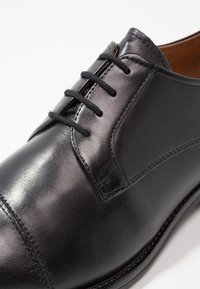 Chaussure habillée en cuir noir avec une finition lisse, dotée de cinq œillets, de lacets noirs et de détails de couture minimalistes. Design classique à bout ovale.