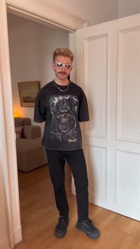 Ung mann med kort blond hår og bart iført sølvfargede solbriller, en svart t-skjorte med hodeskallemotiv, svarte bukser og svarte sko innendørs.