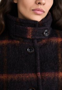 Donna che indossa un cappotto di lana scuro con un motivo a quadri arancione e marrone, colletto alto con bottoni, e grandi bottoni neri, viso parzialmente visibile.