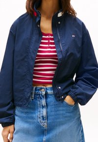 Mörkblå lätt jacka med ståkrage, tryckknappsstängning och elastiska ärmar. Bärs över en röd randig croptop och jeanskjol.