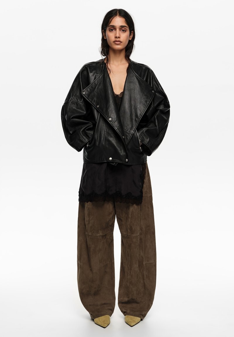 Naine seisab, kandes liiga suurte mõõtudega musta nahktagi, musta pitsist topi, laia pruuni pükstid ja teravate beežide kingadega valgel taustal.