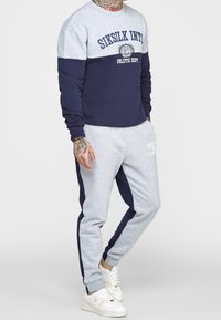 Felpa grigia e blu navy con la scritta "SIKSILK INTL" stampata sul davanti, abbinata a pantaloni grigi con strisce laterali blu navy. Sneakers bianche.