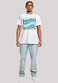 Weißes Baumwoll-T-Shirt mit einem sich wiederholenden "SUPERDAD"-Textdesign in verschiedenen Blau- und Grüntönen. Kombiniert mit hellblauen Jeans und weißen Sneakern.