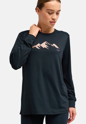 Femme portant un haut noir à manches longues avec un motif beige de montagne sur la poitrine, regardant légèrement vers le bas, une main près du cou.