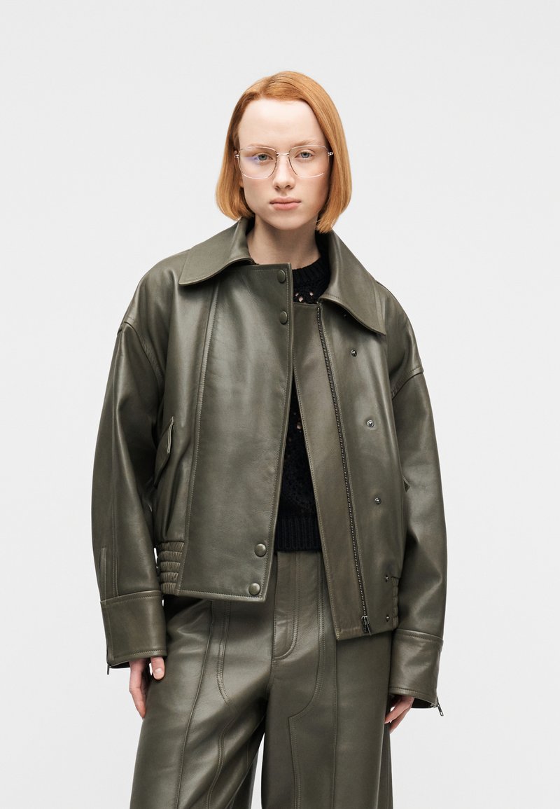 Νεαρό άτομο με κοντά κόκκινα μαλλι ά, φοράει διαφανή γυαλιά, υπερμεγέθη ελαιώδη δερμάτινο μπουφάν, ταιριαστά παντελόνια και μαύρο πλεκτό τοπ σε απλό φόντο.