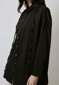 Black button-up shirt with a collar, featuring vertical line details and decorative black ribbons along the sides. Long sleeves and smooth texture.