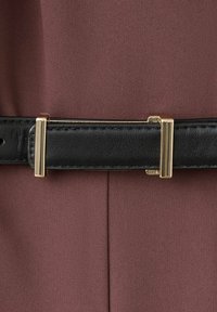 Ceinture en cuir noir avec boucle rectangulaire dorée et passants portée sur un vêtement en tissu brun texturé.