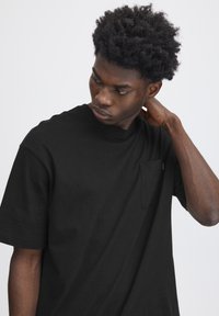 Tricou negru din bumbac, cu guler rotund și mâneci scurte. Prezintă o mică buzunar cusut pe piept. Materialul pare neted, având o croială lejeră.