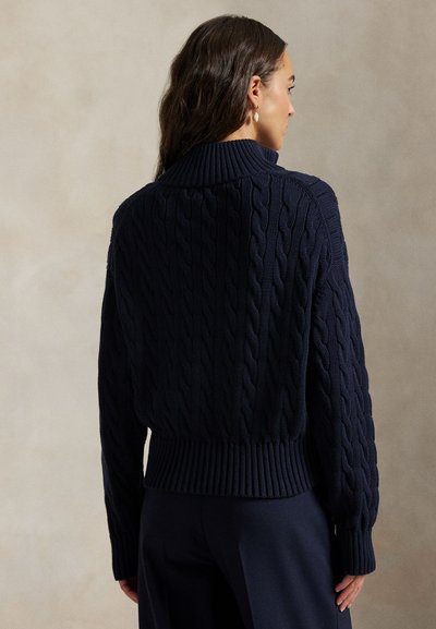 Mujer con cabello oscuro que lleva un suéter de punto trenzado azul marino y pantalones a juego, vista desde atrás sobre un fondo neutro.