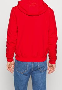 Rød hættejakke med elastiske manchetter og kant. Glat stof, enkelt design. Båret med blå denim jeans med almindelige lommer.