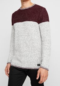 Pull en maille à manches longues présentant une partie supérieure bordeaux et une partie inférieure grise texturée, avec un col rond et des accents côtelés.