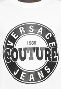 Balta medvilninė marškinėliai su dideliu juodu grafiniu atspaudu, kuriame yra tekstas "VERSACE COUTURE JEANS" ir "1988" apvalioje kompozicijoje.