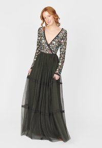 Vestido largo de un verde oscuro, con un corsé de manga larga con estampado floral. Presenta un escote en V y una falda en capas, transparente, con textura de puntos y acentos de encaje.