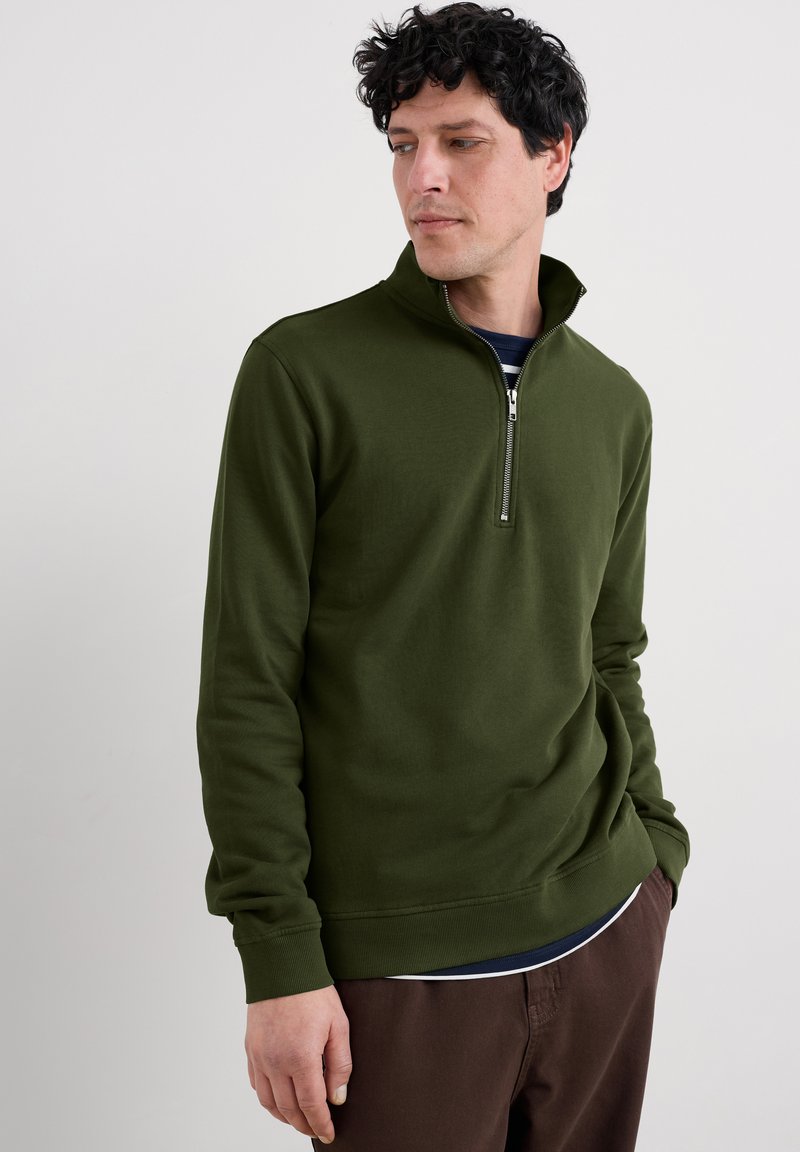 Hombre vistiendo un suéter de cuello semi alto en verde oliva, con una camisa a rayas en azul marino debajo y pantalones marrones, de pie frente a un fondo gris claro.