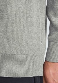 Siv v knitted pulover z teksturiranim vzorcem, rebrastimi manšeti in robom ter majhno oznako blagovne znamke ob strani.