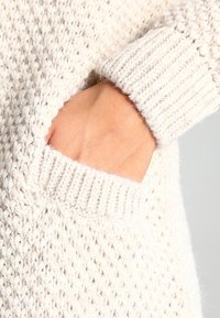 Stickad tröja med en texturerad, krämfärgad tyg och en ribbad mudd. En hand är placerad i en sidoficka.