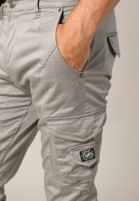 Man met lichtgrijze cargobroek en hand in de voorzak, met gedetailleerde stiksels en een zwart "Declure" label op de zijzak.