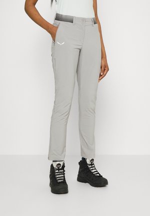 Personne portant un pantalon gris clair ajusté avec des poches latérales et des chaussures de randonnée noires à lacets blancs, debout devant un fond uni.