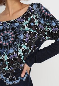 Donna che indossa un top a maniche lunghe blu navy con intricati motivi geometrici e floreali in blu, rosa, viola e turchese, con una mano appoggiata sui fianchi.