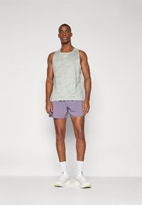 Débardeur vert clair avec un motif de vagues, associé à un short violet. Le modèle porte des baskets blanches avec des accents verts et des chaussettes blanches arrivant aux chevilles.