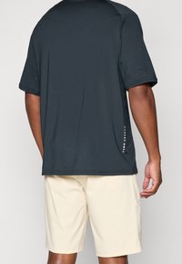 Chemise noire à manches courtes avec une texture lisse, arborant un détail de logo discret sur le côté, associée à un short beige clair.
