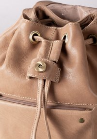 Light brown leather drawstring backpack with brass hardware, a front zipper pocket, and contrasting stitching details. Soft, textured finish.