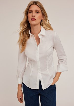 Chemise blanche à boutonnières avec un design ajusté, manches longues retroussées au niveau des poignets et texture de tissu lisse. Détails incluent un col classique et un ourlet arrondi.