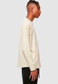 Dlhý rukáv, svetlo béžové tričko vyrobené z bavlny, s voľným strihom, okrúhlym výstrihom a minimálnymi švovými detailmi.