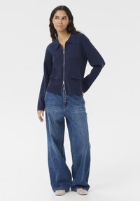 Marinefarbener Reißverschluss-Strickcardigan mit spitzem Kragen, zwei Fronttaschen und langen Ärmeln, kombiniert mit weit geschnittenen blauen Jeans und hellen Schuhen.