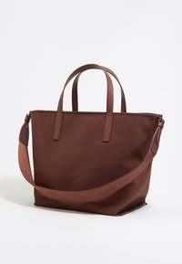 Bolso tote de cuero marrón con asas dobles y una correa de hombro desmontable. Textura suave con un sutil detalle de logo en la correa.