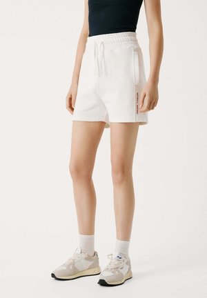 Pantalones cortos de algodón blancos con una cinturilla elástica y cordón, que cuentan con bolsillos laterales y un logo rojo de "Red Bull Racing" en la pierna izquierda.