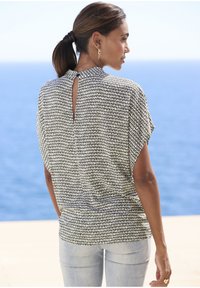 Top a maniche corte, con motivo in bianco e nero e dettaglio a chiave sul retro, abbinato a jeans azzurri chiari, sullo sfondo di un paesaggio marino.