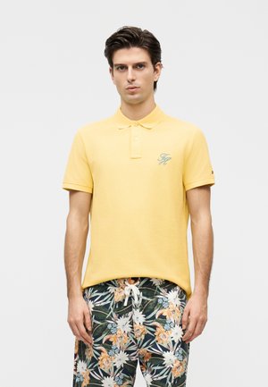 Νεαρός άνδρας που φοράει κίτρινο πόλο πουκάμισο και πράσινα σορτς με λουλουδάτο σχέδιο και κορδόνι, στέκεται μπροστά σε απλό φόντο.