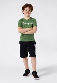 Camiseta Champion verde de mangas curtas com logótipo branco, combinada com calções pretos com texto branco. Ténis desportivos pretos com detalhes em laranja.