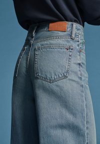 Крупен план на човек, носещ светлосини дънки Tommy Hilfiger с видими задни джобове и кожена маркова етикета на колана.