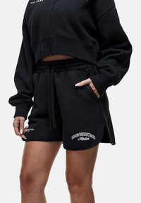 Felpa nera corta e pantaloni corti coordinati in tessuto morbido. I pantaloni presentano una vita elastica, tasche e spacchi laterali con dettaglio del marchio.