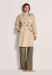 Vrouw in een beige trenchcoat met riem over een roze coltrui, bruine broek met wijde pijpen en huidkleurige hakken, staand tegen een effen muur.