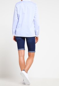 Camisa de manga larga azul claro con corte recto, combinada con pantalones cortos de mezclilla oscuros hasta la rodilla y zapatillas blancas. Texturas suaves, ajuste casual.