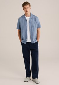 Camisa de botón de manga corta en azul claro, camiseta blanca por debajo, pantalones azul marino y zapatos grises. Cuenta con un bolsillo en el pecho y un corte relajado.
