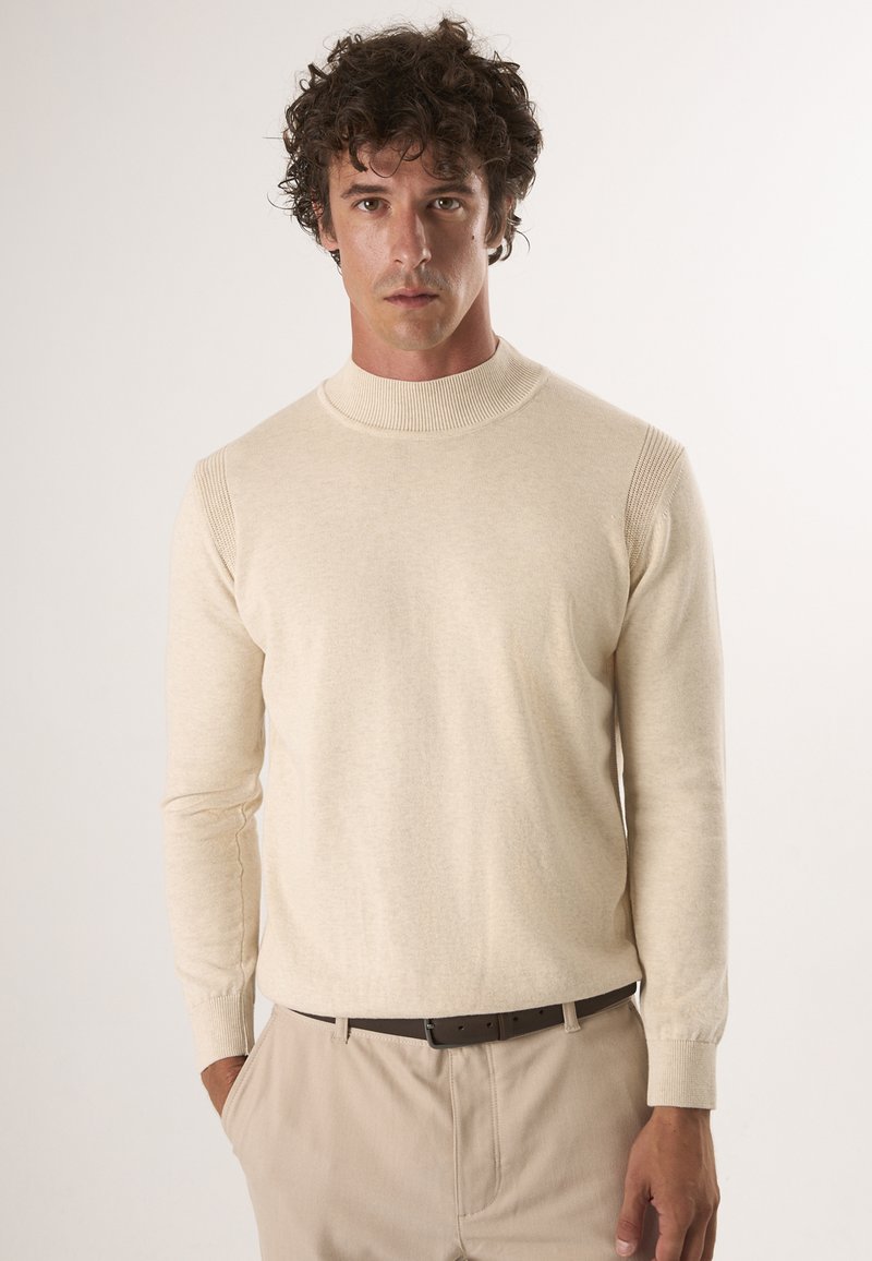 Beige gebreid trui met een hoge nek, lange mouwen, ribbelaccenten op de schouders en een gladde textuur, gecombineerd met lichtgekleurde broek.