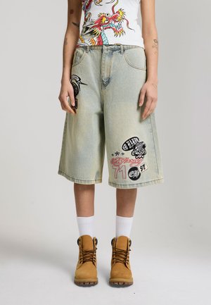 Person trägt hellblaue Jeans-Shorts bis zu den Knien mit grafischen Aufnähern, weiße Sportsocken, hellbraune Schnürstiefel und ein weißes ärmelloses Oberteil mit einem Drachendesign.