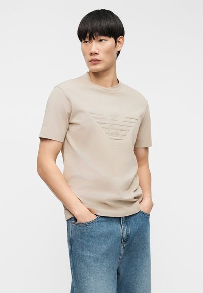 Φωτεινή μπεζ βαμβακερή μπλούζα με ανάγλυφο σχέδιο λογότυπου, στρογγυλή λαιμόκοψη και κοντά μανίκια, σε συνδυασμό με άνετο μπλε τζιν παντελόνι.