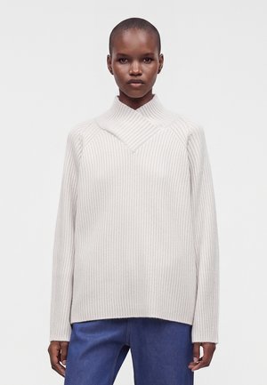 Personne à la peau foncée aux cheveux courts portant un pull léger en maille côtelée et un pantalon bleu, debout sur un fond blanc uni.