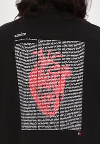 Juodas marškinėliai su raudono širdies grafika ir tekstiniu dizainu. Tekstas apibūdina "amore" įvairiais formatais, su išraiškinga širdies iliustracija.