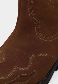 Botte de cowboy en daim marron avec coutures blanches décoratives et semelle noire, montrant un gros plan du côté supérieur et des détails du motif complexe.
