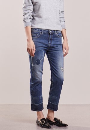 Persoon draagt cropped blauwe spijkerbroek met patch en versleten details, lichtgrijs top met lange mouwen en zwarte instapschoenen met gouden gespen.