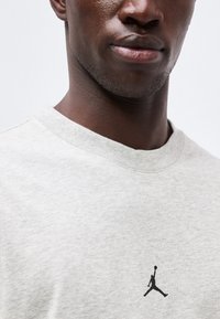 Κοντινό πλάνο ενός ατόμου που φοράει ανοιχτό γκρι πουκάμισο με μικρό μαύρο λογότυπο Jumpman κοντά στην περιοχή του στήθους.